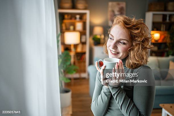 lächelnde junge frau, die zu hause eine tasse kaffee trinkt - kräutertee stock-fotos und bilder