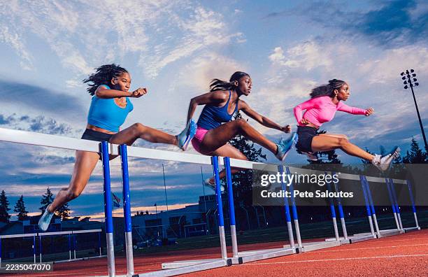 three athletes in vibrant sportswear jumping hurdles on a track with a scenic sky backdrop. usa - agilidad fotografías e imágenes de stock