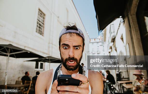 a man reacts comically with shock while checking his phone in a bustling outdoor place in a historic city - shock stock pictures, royalty-free photos & images
