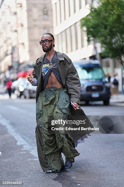 Guest wears short closely cropped hair, oversized brown-tinted aviator sunglasses with dark frames, a silver cross pendant necklace and multiple...