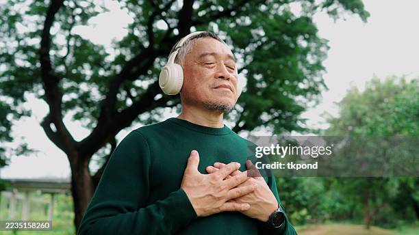 elderly asian man meditating under a tree with wireless headphones, calm and refreshed - welzijn stockfoto's en -beelden