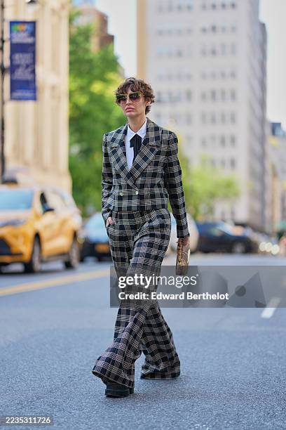 Irina Kro wears short tousled brown hair with lighter highlights, oversized translucent green cat-eye sunglasses with dark lenses, small gold hoop...