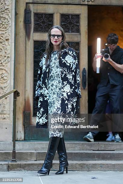 Jordan Roth wears long center-parted brown hair worn loose with soft waves, black rectangular sunglasses, a black embroidered coat with white floral...