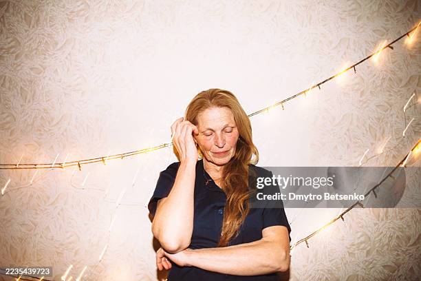 portrait of senior woman standing by the wall with christmas lights - hand in hair stock pictures, royalty-free photos & images