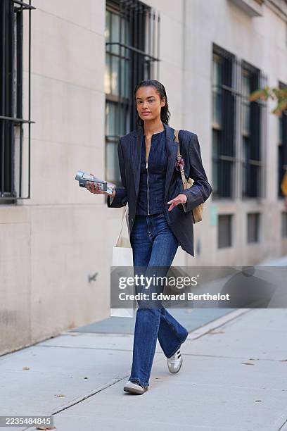 Guest wears slicked-back wet-look dark brown hair, a tan leather shoulder bag with a single strap, gold-tone hardware and a purple floral charm, a...