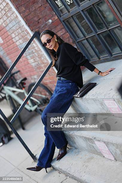 Guest wears straight dark brown hair worn down with a black headband, small oval black-framed sunglasses with dark lenses, gold-tone ribbed earrings...