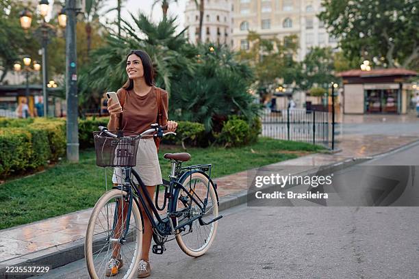 mujer joven usando un teléfono inteligente mientras está de pie con su bicicleta en la ciudad - navegación fotografías e imágenes de stock