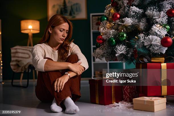 sad woman sitting on floor by christmas tree with presents - december stockfoto's en -beelden