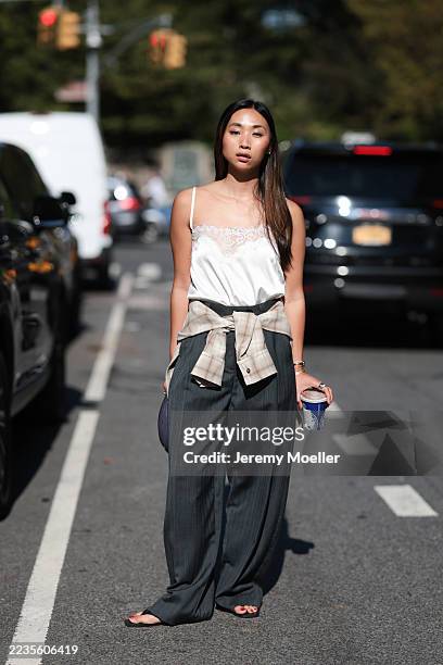 Fashion week guest is seen wearing a white satin camisole with lace detailing at the neckline, paired with dark gray wide-leg pants featuring subtle...