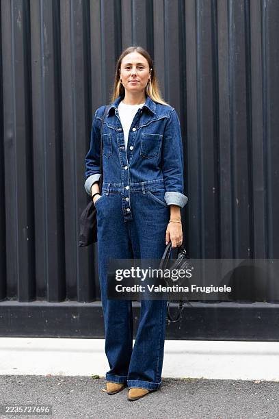 Person wears gold hoop earrings, a white t-shirt, a black tote bag, a black leather studded cross-body bag, a denim jumpsuit and beige suede boots...