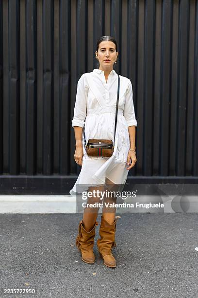 Person wears gold pendant earrings, a white long sleeved short dress, a Gucci suede cross-body mini bag and beige suede cow boy boots with fringes...
