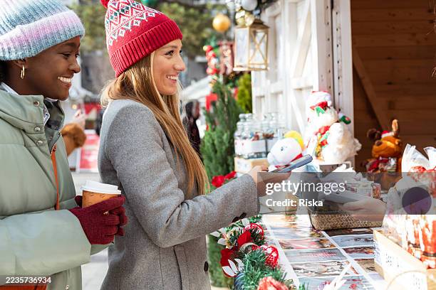 two friends are shopping at a christmas market booth during the holiday season - mobile payment stock pictures, royalty-free photos & images