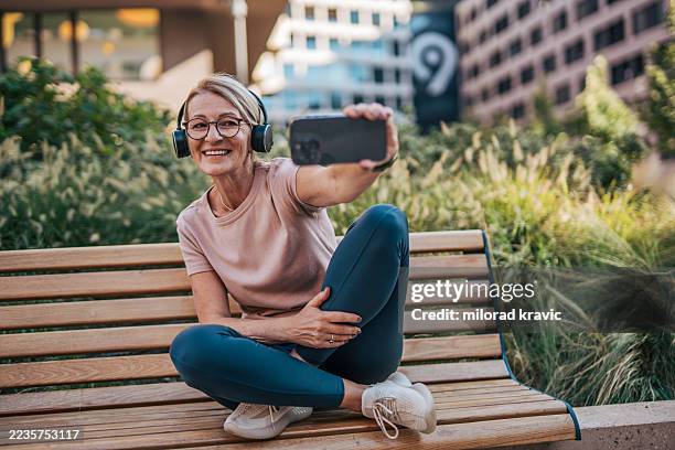 senior adult woman taking a selfie while listening music on a bench - park bench stock pictures, royalty-free photos & images