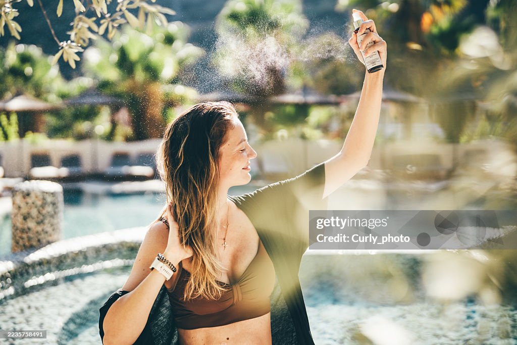 A woman sprays her hair with protective spray to prevent dryness while sitting by a turquoise pool.