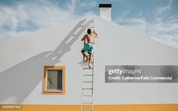conceptual image of a topless man in swimming shorts climbing the side of a white house on a ladder, and casting a long shadow - escalera plegable fotografías e imágenes de stock