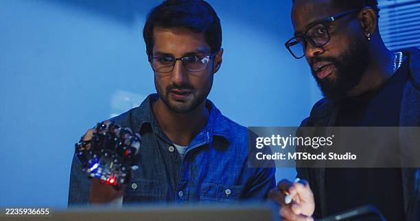 black and hispanic male engineers programmers work together on robot hand prototype, use computer control robotic arm circuit in tech office night. software developer, artificial intelligence. - robotic prosthetic stock pictures, royalty-free photos & images