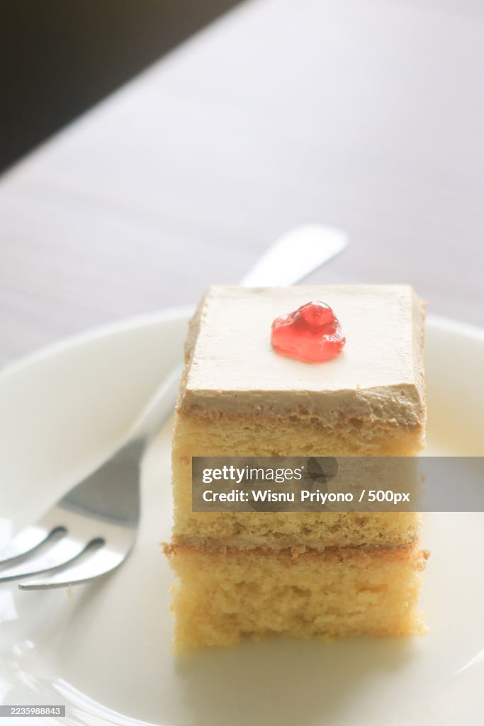Close-up of a square slice of cake with frosting and a cherry on a white plate with a fork