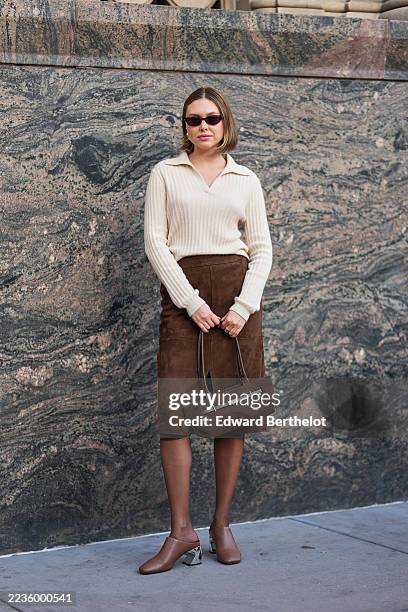 Guest wears a chin-length light brown bob with a center part, small rectangular dark brown sunglasses, small gold hoop earrings, a brown suede...