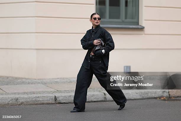 Sophia Geiss seen wearing narrow oval 90s-inspired black sunglasses, a black denim shirt, slightly oversized with structured shoulders and chest...
