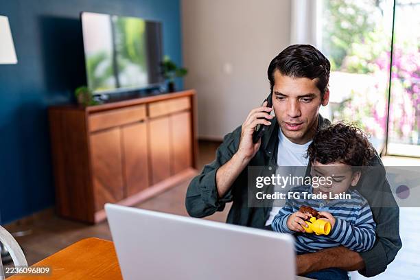 young man working and talking on mobile phone while holding his baby son at home - house husband stock pictures, royalty-free photos & images