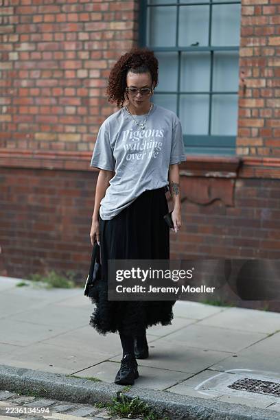 Guest wears a loose, grey graphic T-shirt with the slogan “Pigeons watching over me,” paired with a black midi skirt featuring voluminous ruffled...