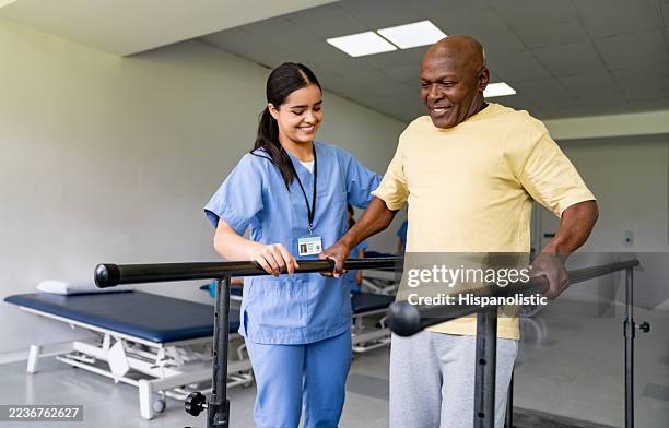 physical therapist helping a senior man walking on bars at a rehabilitation center - therapie stockfoto's en -beelden