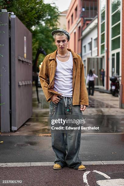 Guest wears short bleached blonde hair topped with a grey baker boy cap, a thick silver chain necklace and a small silver hoop earring with multiple...