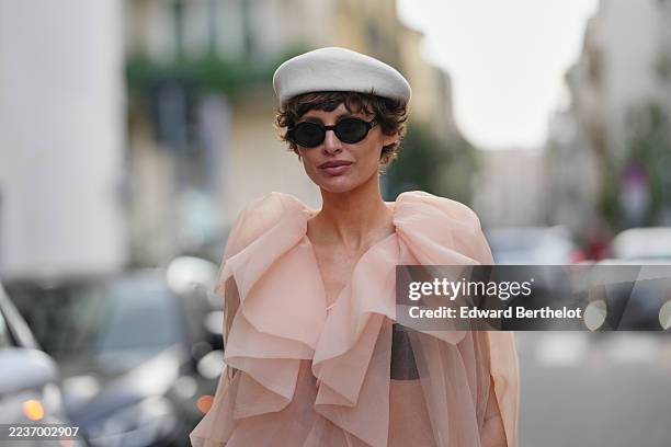 Irina Kro wears a short tousled dark brown curly hairstyle, a white felt beret hat, black oval sunglasses, a layered pale peach sheer organza cape...