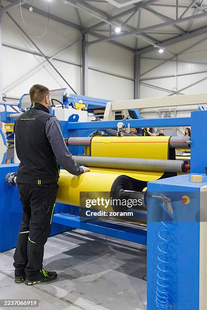 worker operating yellow rollers on large blue industrial printing machine in manufacturing plant - rolling paper stock pictures, royalty-free photos & images