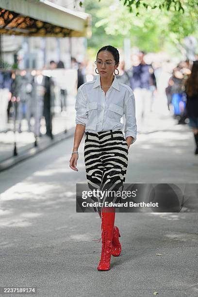 Yoyo Cao wears a slicked-back low bun hairstyle, dark brown, transparent rectangular glasses, large gold hoop earrings, a silver chain necklace, a...