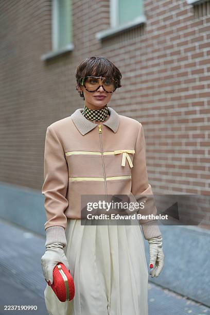 Irina Kro wears a short dark brown cropped haircut with fringe and a slight center part, oversized brown-tinted sunglasses, small gold hoop earrings,...