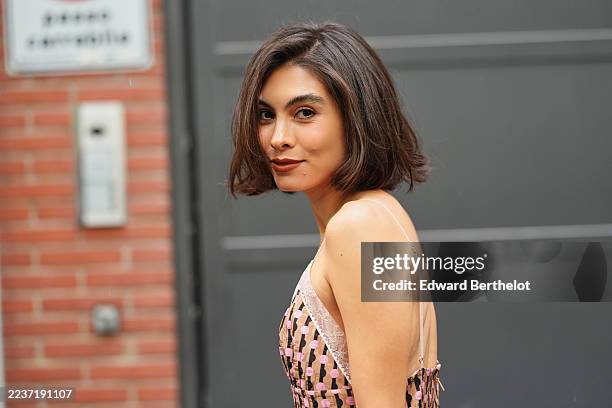 Fer Millan Delaroiere wears a chin-length dark brown bob with a side part, a thin gold chain necklace, a lace-trimmed spaghetti-strap dress with pale...