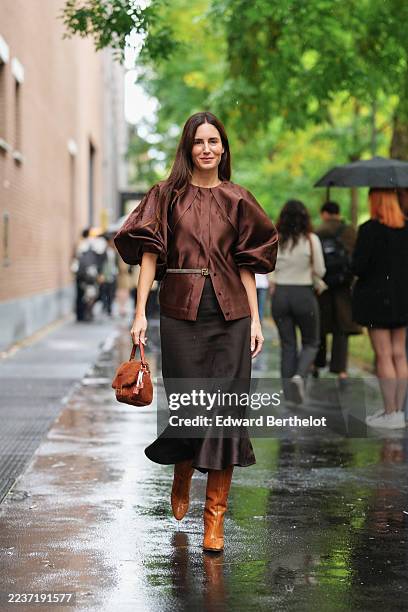 Gala Gonzalez wears long straight dark brown hair with a center part, a gold ring on the hand, a brown shearling clutch bag with a leather strap...