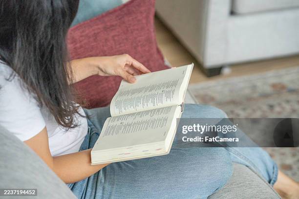 cozy moment of a woman reading a book on a sofa at home, relaxed and focused - paperback stock pictures, royalty-free photos & images