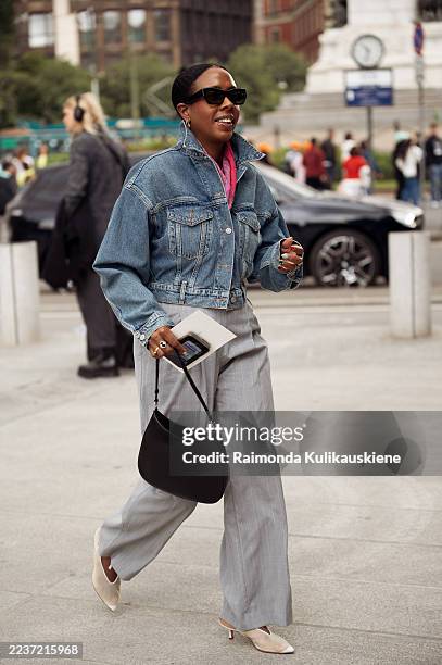 Guest wears a cropped denim jacket layered over a pink scarf with grey pleated wide-leg trousers and white pointed heels and carries a black Prada...