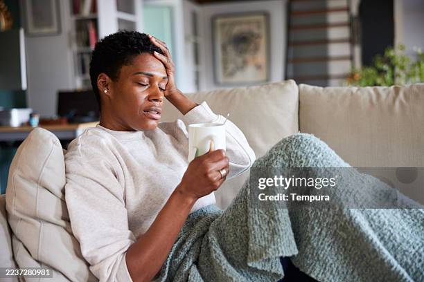 a woman lies on a sofa with her hand on her forehead, holding a mug, and appears to have a headache. - symptom stock pictures, royalty-free photos & images
