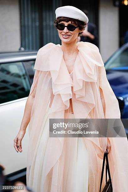 Irina Kro Eicke is seen wearing a layered pale pink chiffon cape with a wide ruffled collar, a classic white beret and vibrant red pointed-toe flats,...