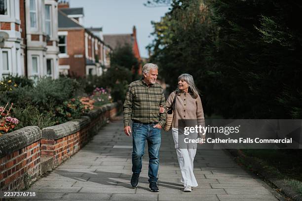 a mature couple walk arm-in-arm down a footpath in a residential district - pensionierung stock-fotos und bilder