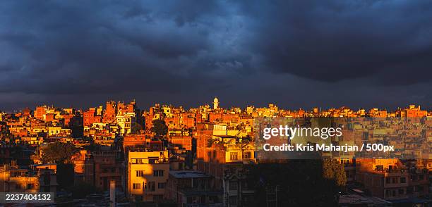 panoramic view of a city skyline with buildings illuminated by golden light against a dramatic, dark sky at sunset - kathmandu nepal stock pictures, royalty-free photos & images