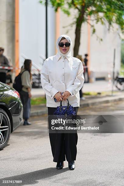 Guest wears a white silk headscarf wrapped close to the face, black angular cat-eye sunglasses, a navy blue fabric top-handle bag from Prada with...