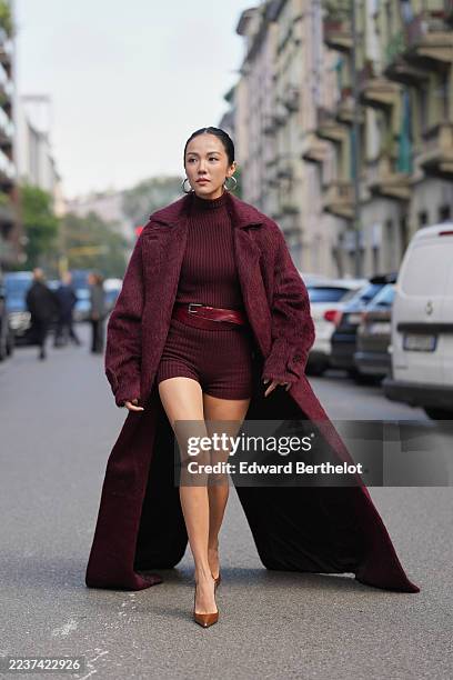 Yoyo Cao wears pulled-back dark brown hair in a low bun with a center part, large silver hoop earrings and silver rings, a long burgundy fur coat...