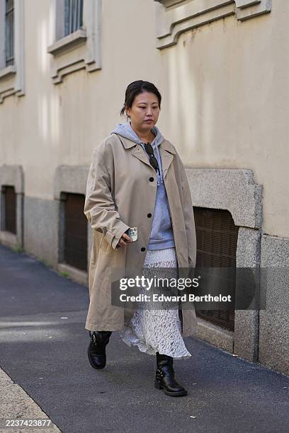 Guest wears dark brown pulled-back hair in a low bun, black sunglasses hanging from the light grey hoodie neckline, a beige long trench coat with...