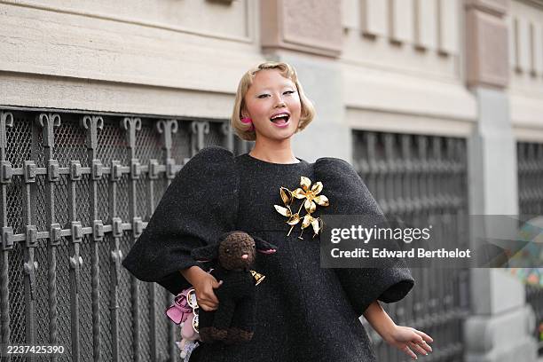 Lin Lin wears a short blonde bob with a soft wave and side part, pink round earrings, a gold-tone large floral brooch, a micro pink bag from Moschino...