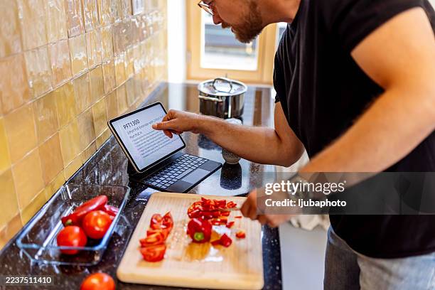 focused cook chops peppers while reading on tablet in modern kitchen with tomatoes - chopped chilli stock pictures, royalty-free photos & images