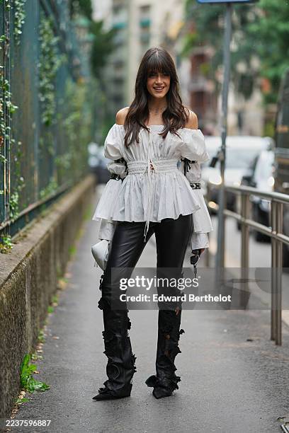 Guest wears long dark brown hair with blunt bangs and no visible part, black rectangular sunglasses, layered silver necklaces, a white leather pouch...