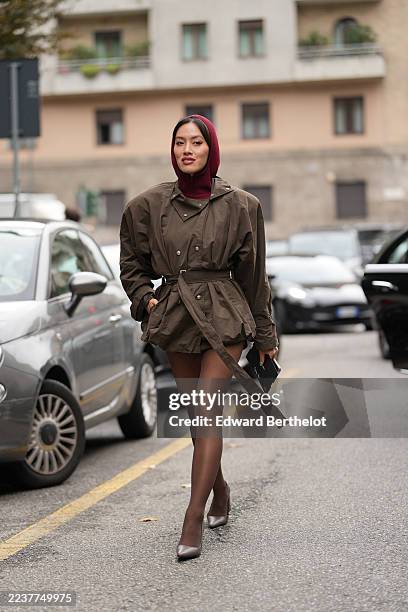 Tiffany Hsu wears straight black chin-length hair with a center part, a burgundy knit balaclava covering the head and neck, a short brown belted...