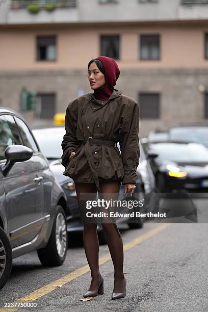 Tiffany Hsu wears straight black chin-length hair with a center part, a burgundy knit balaclava covering the head and neck, a short brown belted...
