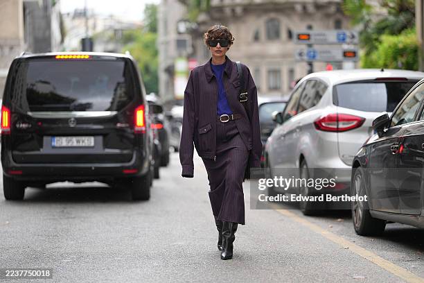 Melanie Kieback wears short tousled brown hair, oversized black sunglasses, a black leather shoulder bag with gold buckle hardware, a purple knit top...
