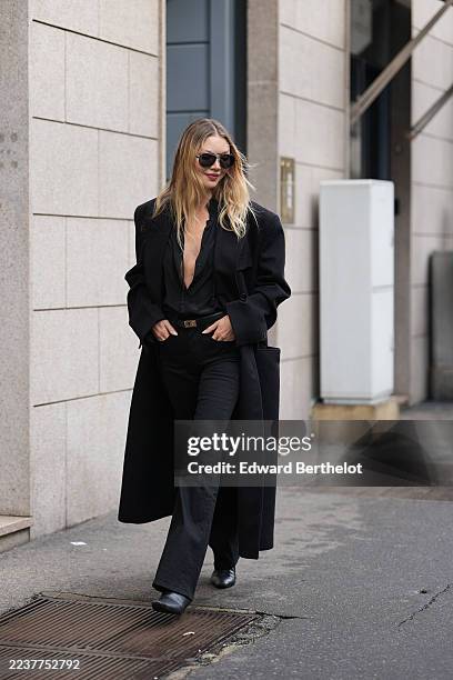 Jessie Andrews wears long blonde wavy hair with a center part, oversized black sunglasses, a long black coat with structured shoulders, a black satin...