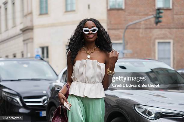 Guest wears long black curly hair with a center part, white cat-eye sunglasses, a chunky gold chain necklace with a round pendant and stacked gold...